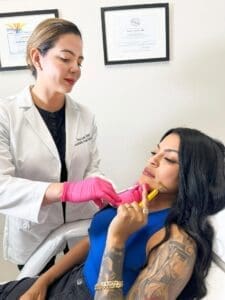 Specialist Wearing A White Coat Black Uniform And Pink Gloves Applies Radiesse Dermal Fillers Injection To A Woman Holding A Facial Roller To Her Face