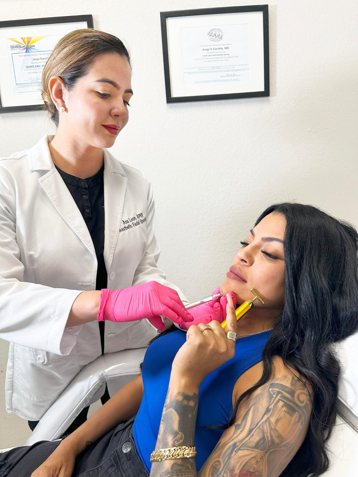 Specialist Wearing A White Coat Black Uniform And Pink Gloves Applies Radiesse Dermal Fillers Injection To A Woman Holding A Facial Roller To Her Face